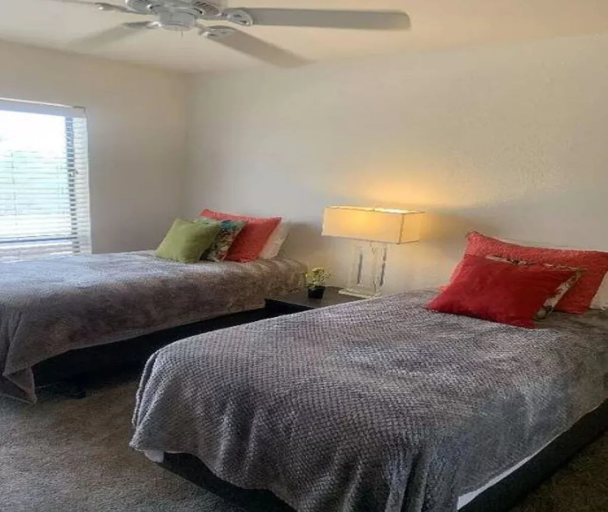 A tranquil bedroom with two twin beds, each adorned with decorative pillows. A standing lamp and window with blinds provide soft natural light, creating a cozy atmosphere ideal for quiet moments or small gatherings. The layout suggests an inviting space for both rest and connection.