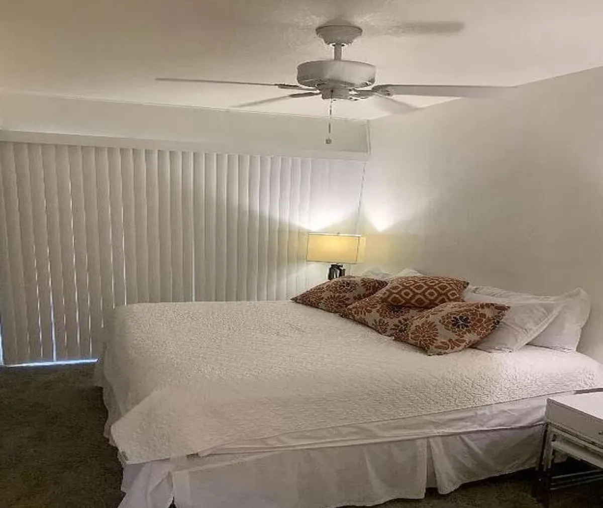 An inviting bedroom scene showcasing a neatly made bed with decorative pillows, a bedside lamp, wall decor, and a ceiling fan, all bathed in natural light. The serene ambiance and simple decor evoke peace and comfort, making it an ideal retreat for unwinding after group activities.