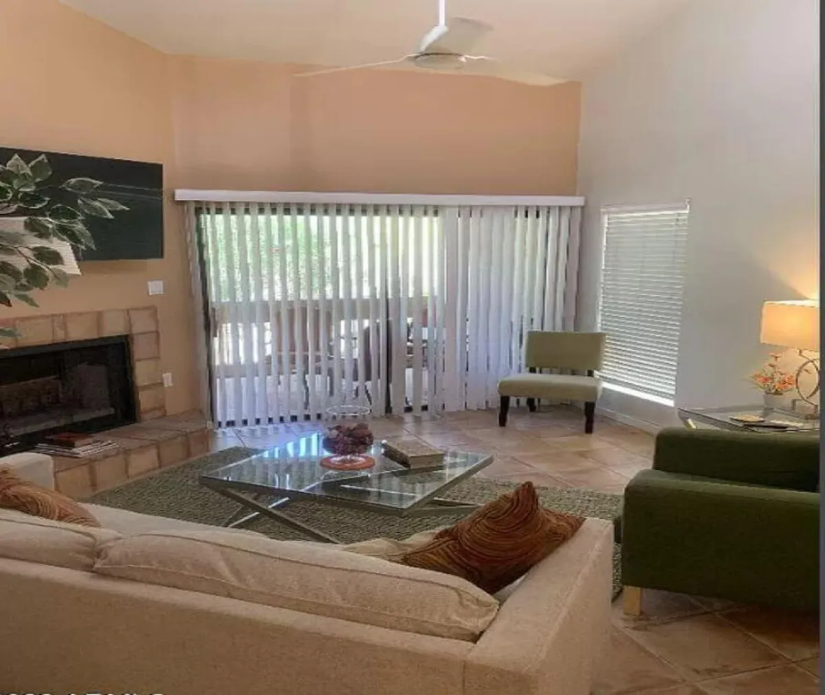 A cozy living room featuring a welcoming fireplace, a soft sofa, green accent chairs, a glass coffee table, and window blinds that let in ample natural light. The arrangement invites conversation and relaxation, and the overall setting feels both inviting and serene.