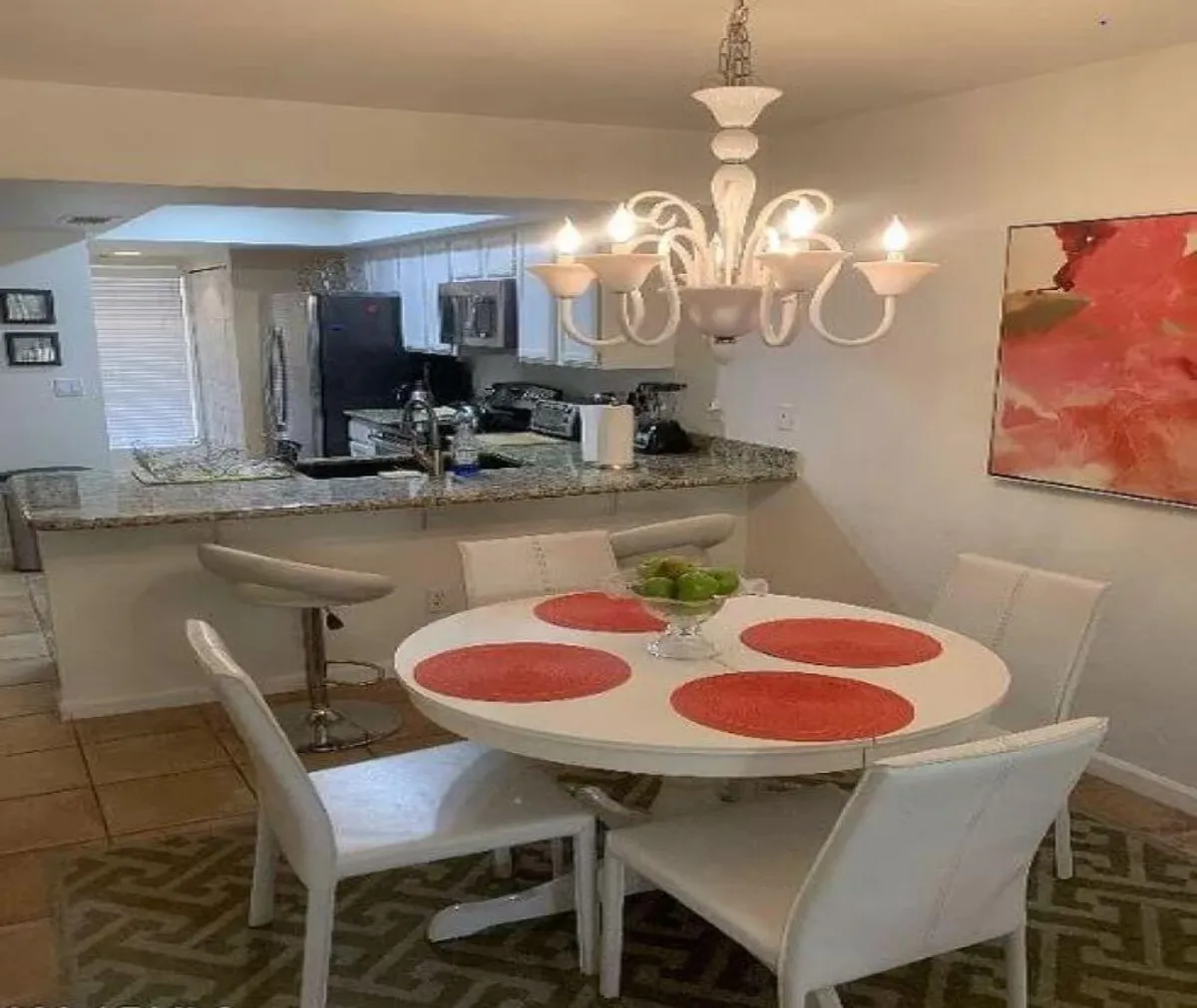 The image showcases a warm and inviting open-plan dining and kitchen area. A round dining table sits under a stylish chandelier, flanked by modern bar stools at a spacious kitchen counter. Abstract wall art adds a contemporary flair, while natural light fills the space, creating a perfect setting for social gatherings or intimate meals.